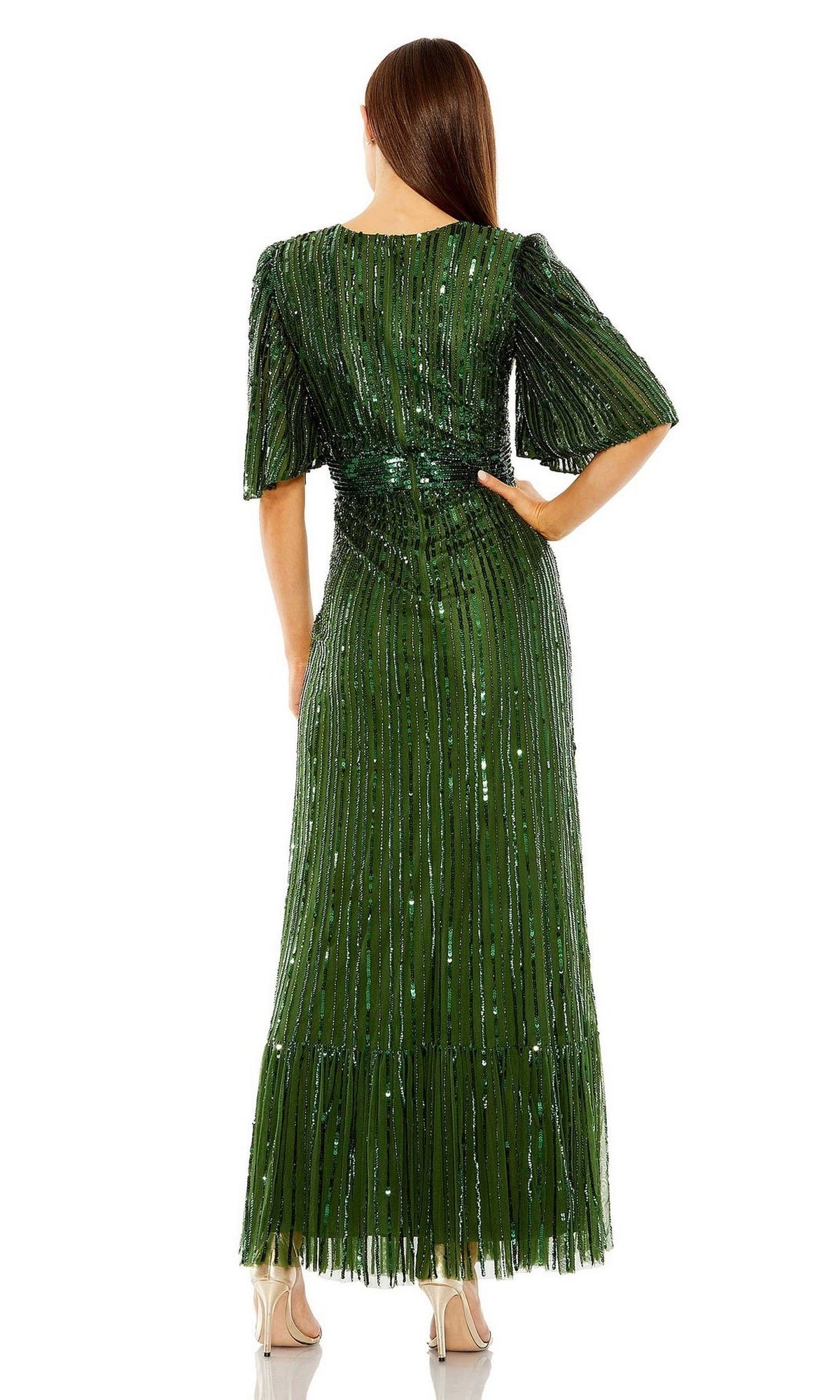 A woman stands with her back to the camera, wearing the Long Formal Dress 23010 by Mac Duggal—an emerald green, floor-length sequin gown with short sleeves and beige heels.